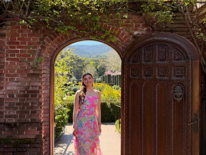 Kelsey Sweetland in a pink floral dress, smiling, framed by a rounded brick archway with a wooden door and a sunlit garden behind her.